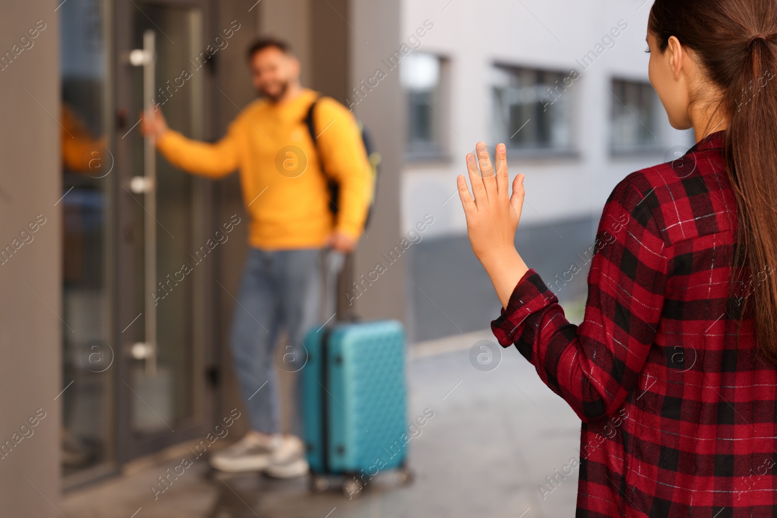 Photo of Long-distance relationship. Woman waving to her boyfriend with luggage near building outdoors