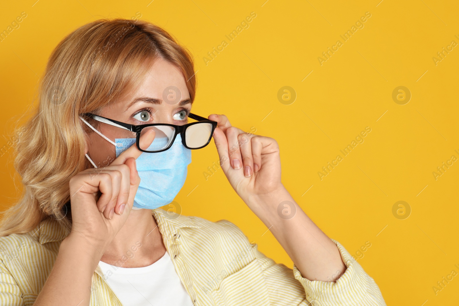 Photo of Woman wiping foggy glasses caused by wearing medical mask on yellow background