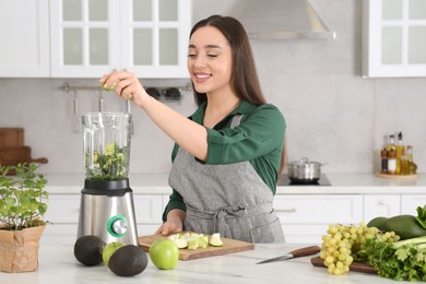 Woman adding apples into blender with ingredients for smoothie at white table in kitchen