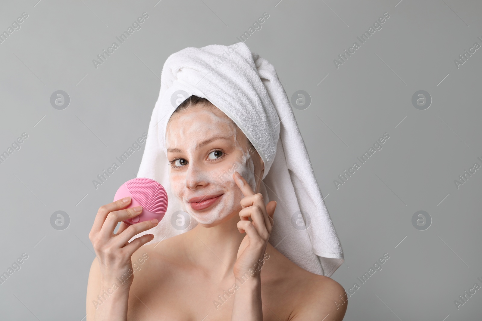 Photo of Young woman washing face with brush and cleansing foam on grey background