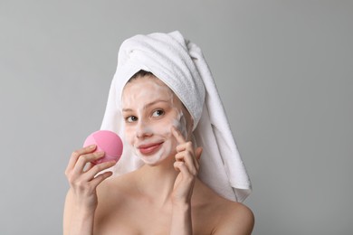 Young woman washing face with brush and cleansing foam on grey background