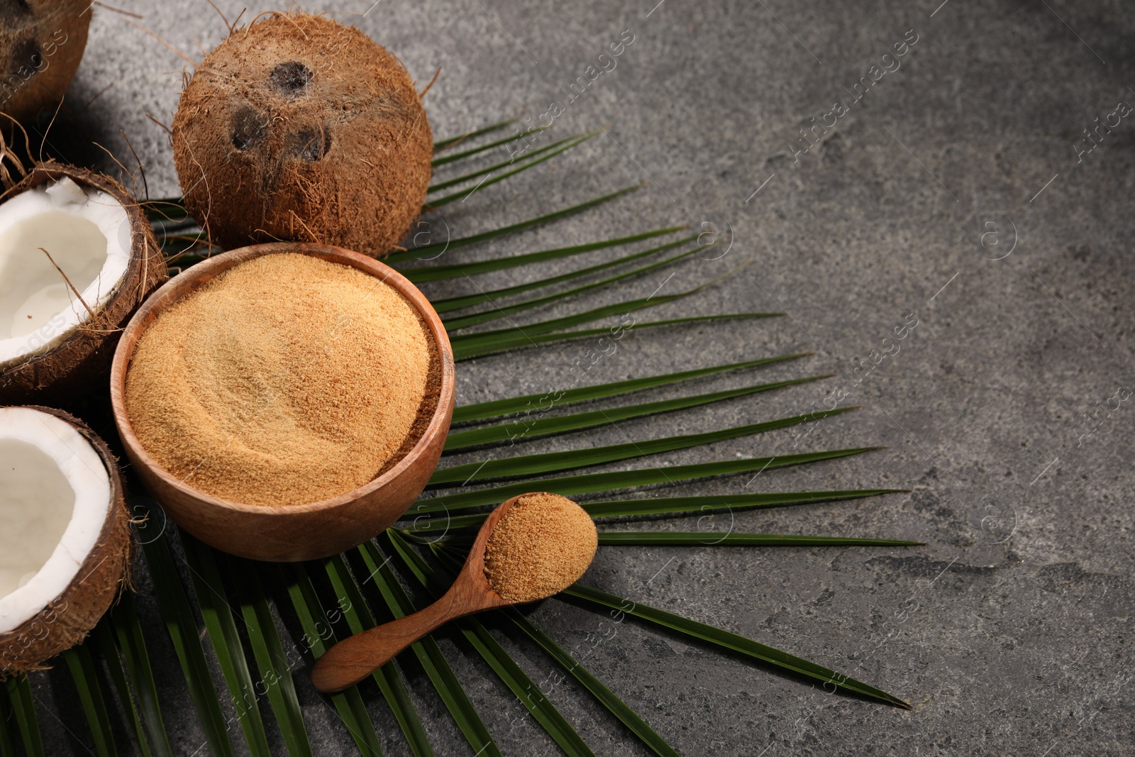 Photo of Coconut sugar in bowl, spoon, palm leaves and fruits on dark textured table, closeup. Space for text