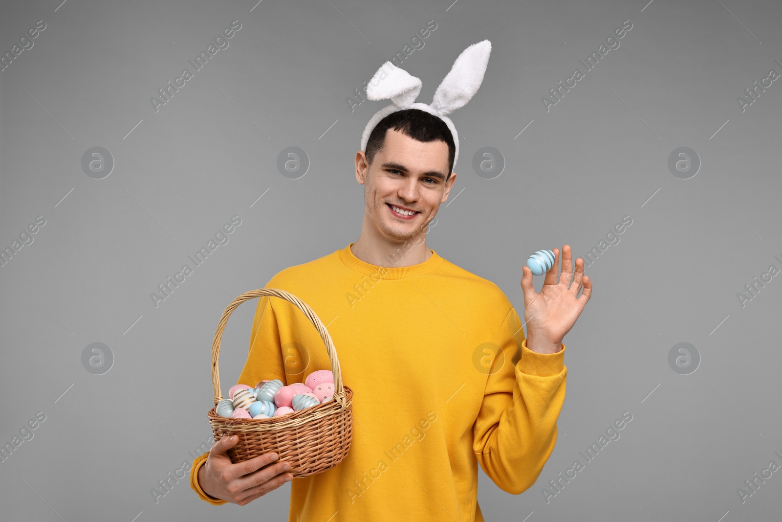 Photo of Easter celebration. Handsome young man with bunny ears and painted eggs on grey background