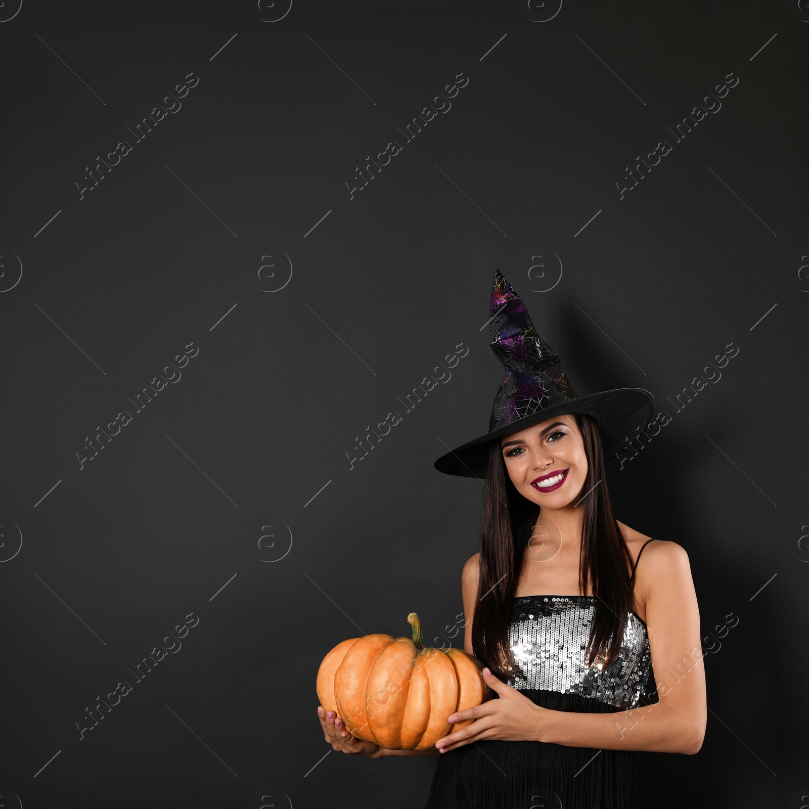 Photo of Beautiful woman wearing witch costume with pumpkin for Halloween party on black background, space for text