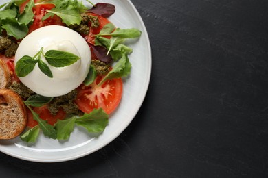 Photo of Delicious burrata salad with tomatoes, arugula and pesto sauce on black table, top view. Space for text