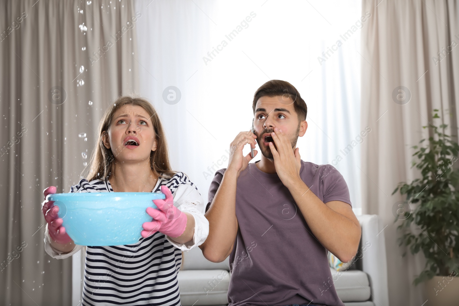 Photo of Emotional young woman collecting water leaking from ceiling while her husband calling plumber in living room