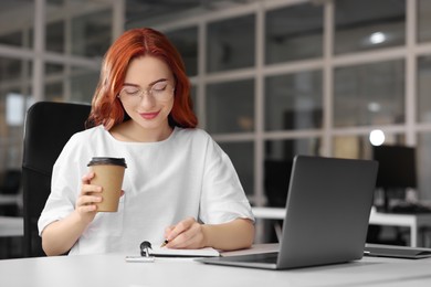 Happy woman with paper cup of coffee taking notes while working on laptop at white desk in office