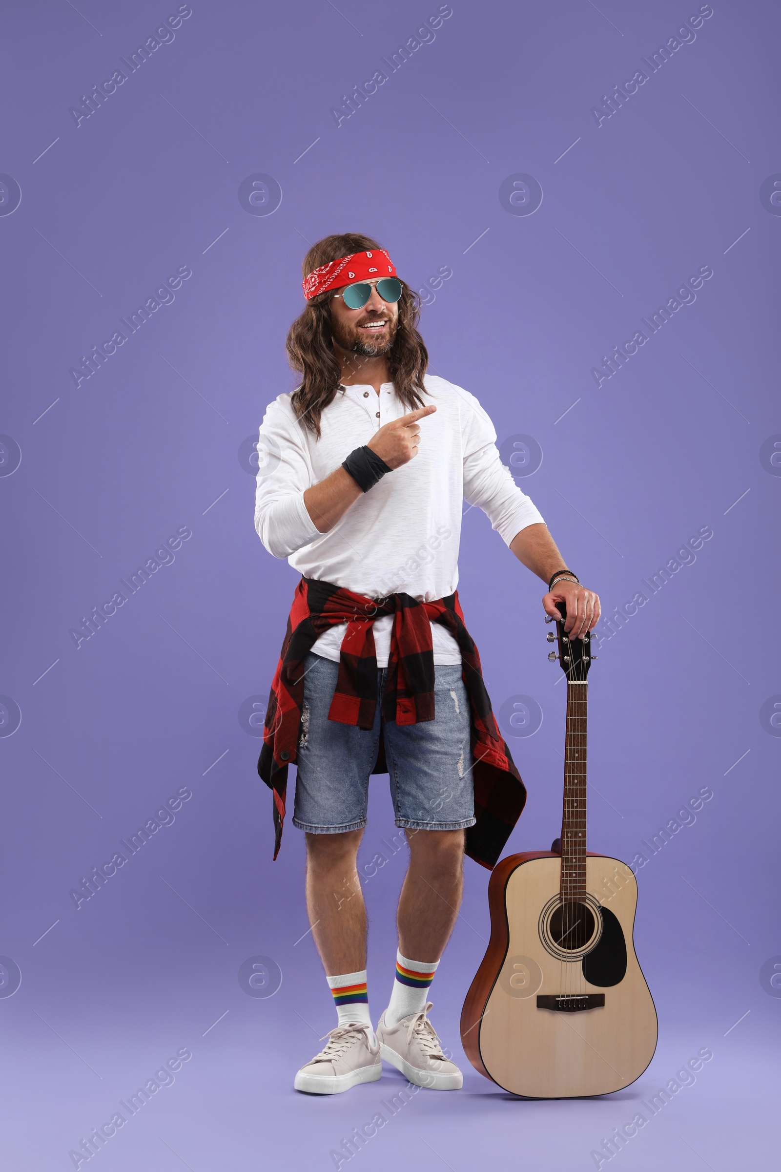 Photo of Stylish hippie man with guitar pointing at something on violet background