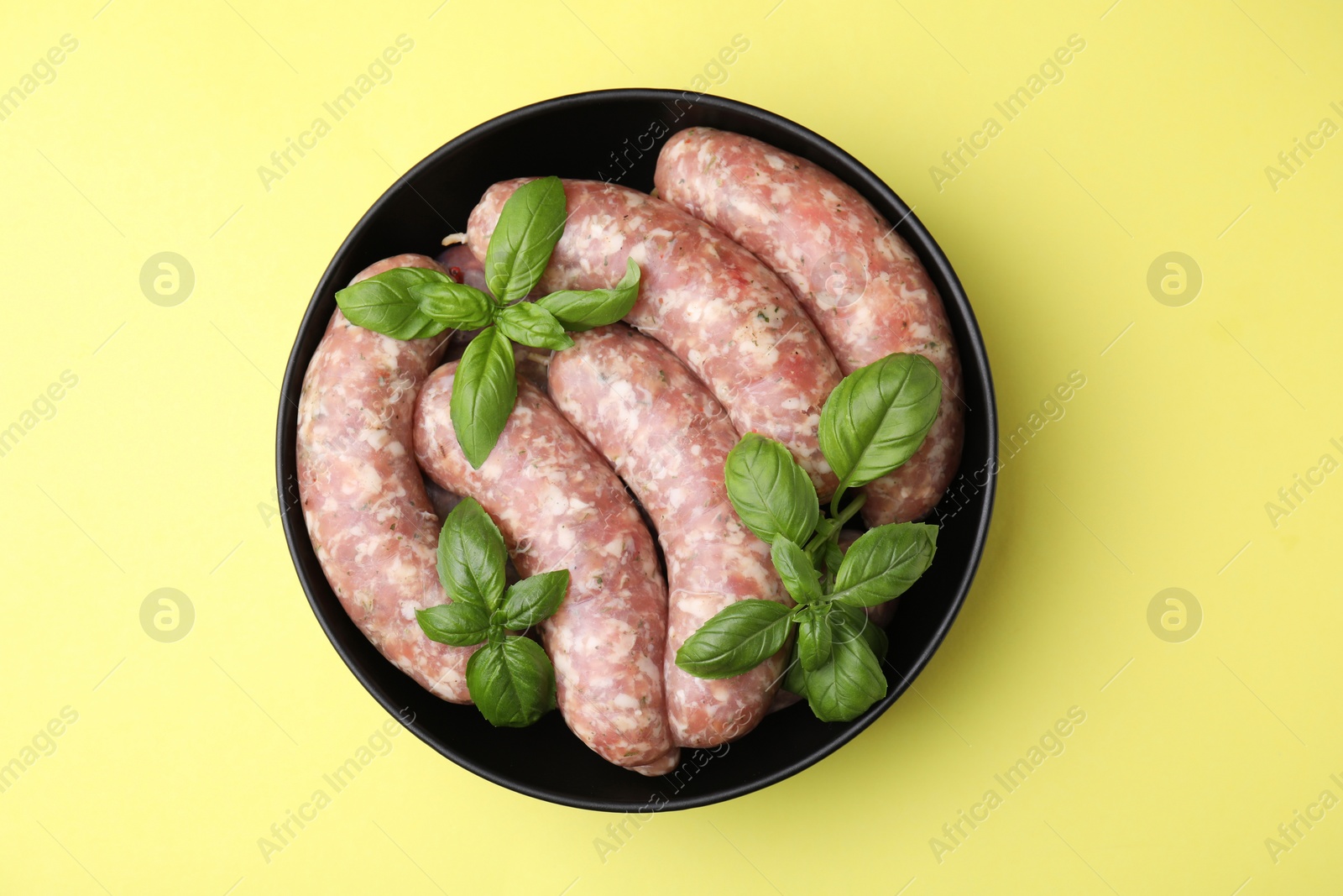 Photo of Raw homemade sausages and basil leaves in bowl on yellow background, top view