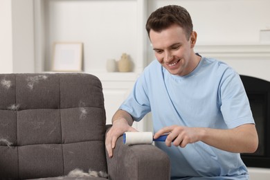 Photo of Pet shedding. Smiling man with lint roller removing dog's hair from armchair at home