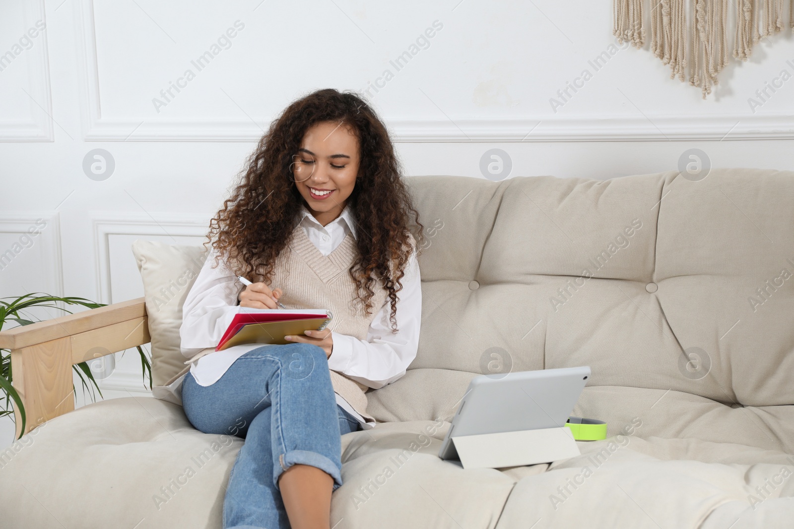 Photo of African American woman with notebook and tablet studying on sofa at home. Distance learning