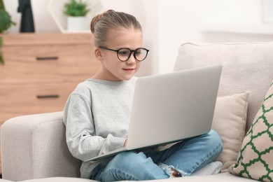 Photo of Little girl in glasses using laptop on sofa indoors