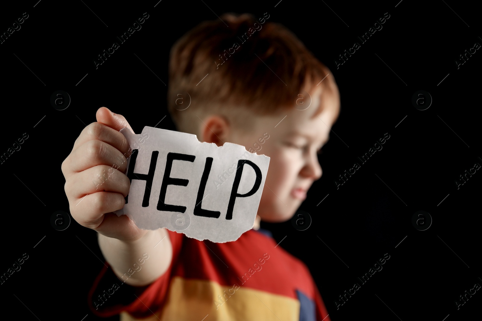 Photo of Little boy holding piece of paper with word Help against black background, focus on hand. Domestic violence concept