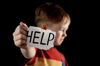 Little boy holding piece of paper with word Help against black background, focus on hand. Domestic violence concept