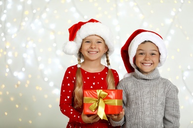 Photo of Happy little children in Santa hats with gift box against blurred festive lights. Christmas celebration