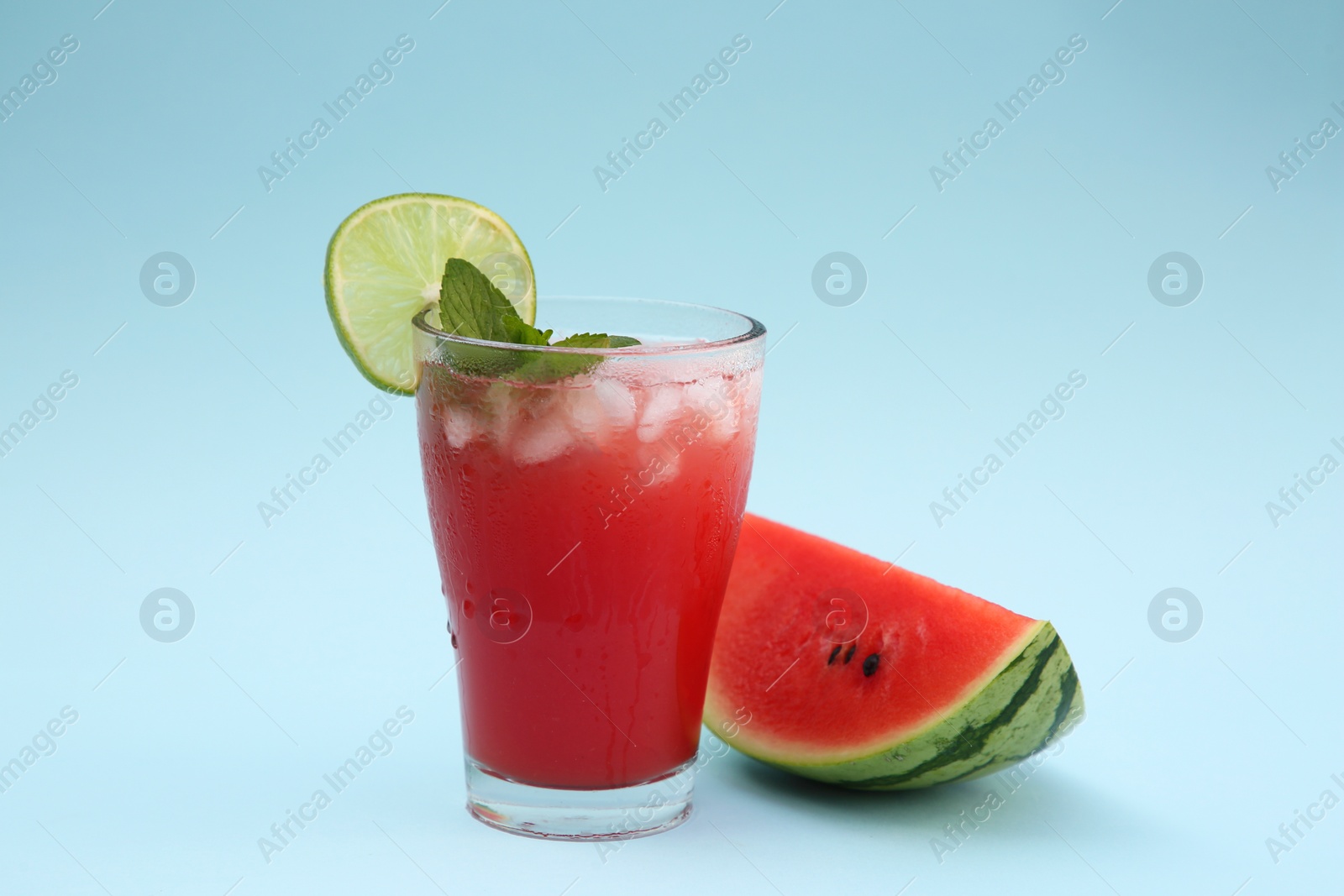 Photo of Glass of delicious drink with lime, ice cubes and cut fresh watermelon on light blue background