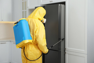 Pest control worker in protective suit spraying insecticide near refrigerator indoors