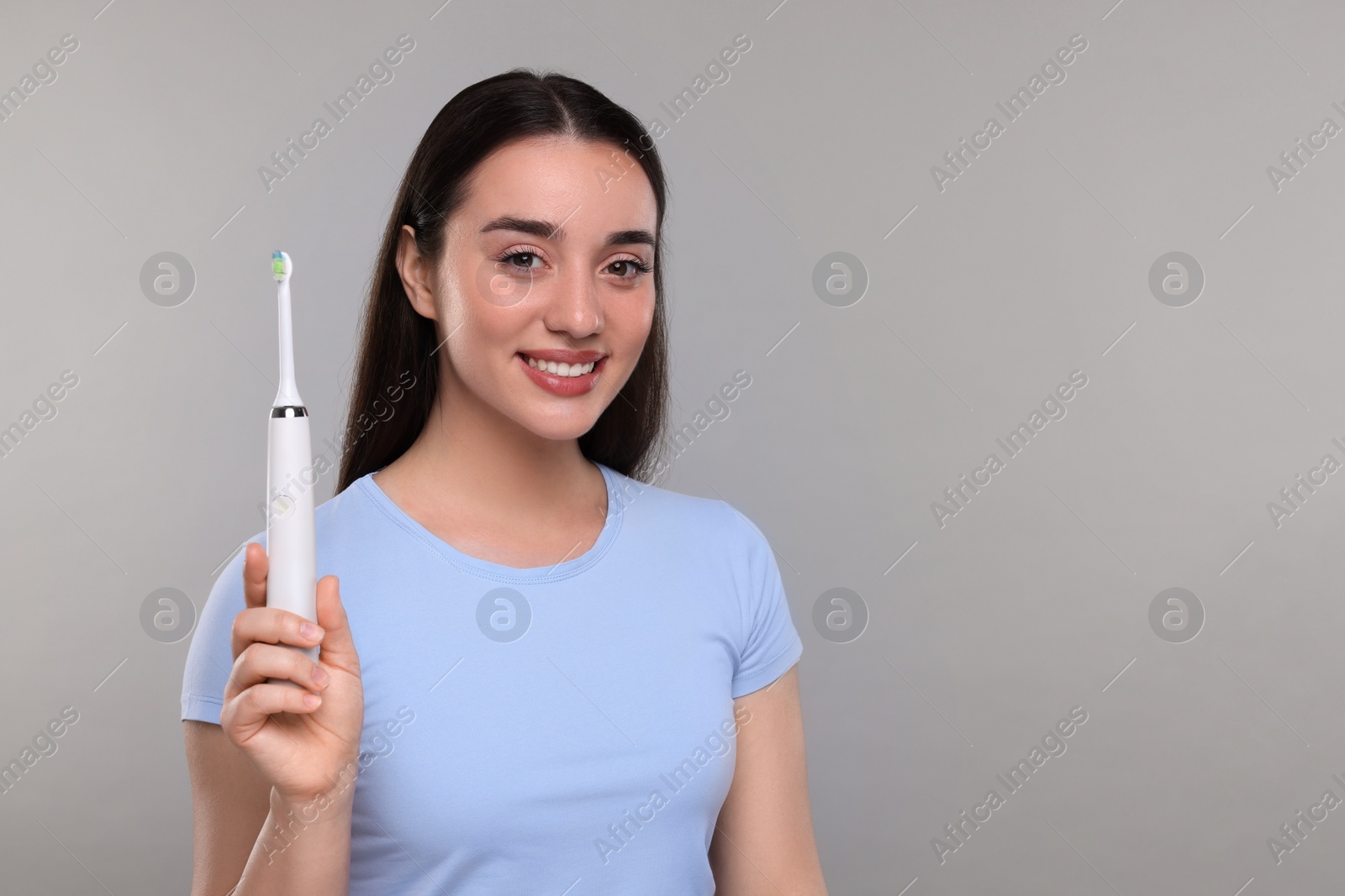 Photo of Happy young woman holding electric toothbrush on light grey background, space for text