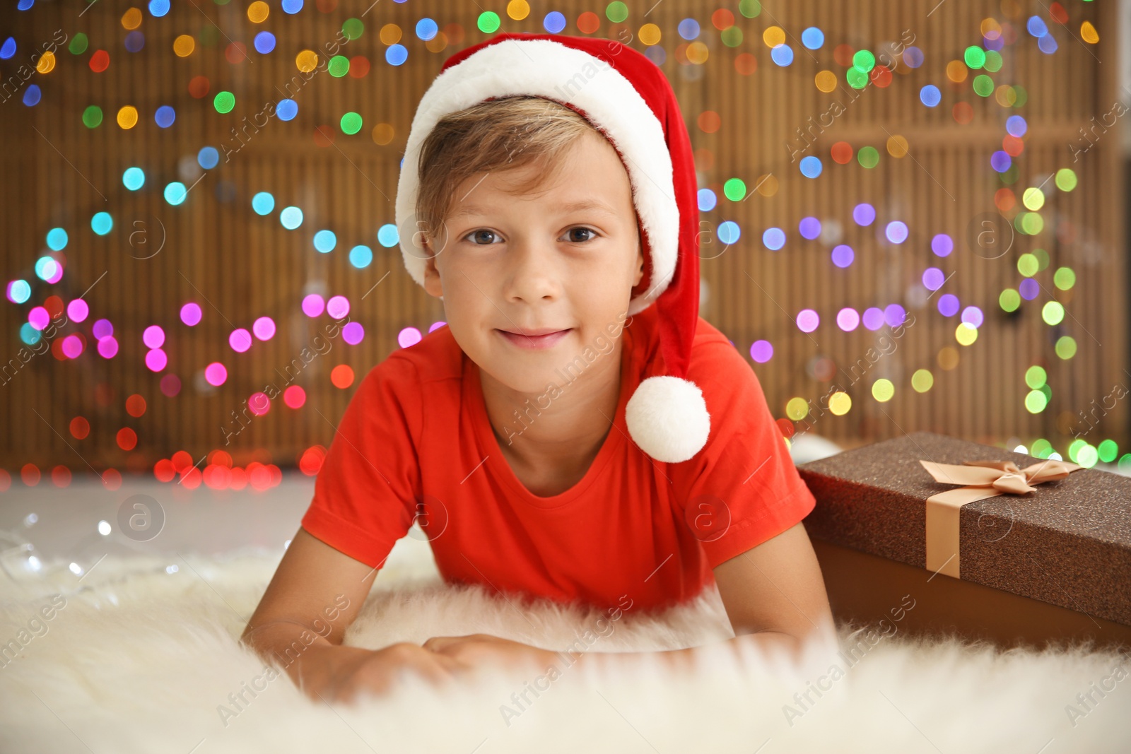 Photo of Cute little child in Santa hat with Christmas gift box lying on floor against blurred lights
