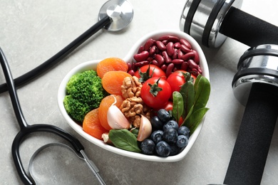Photo of Bowl of products for heart-healthy diet, dumbbells and stethoscope on grey table