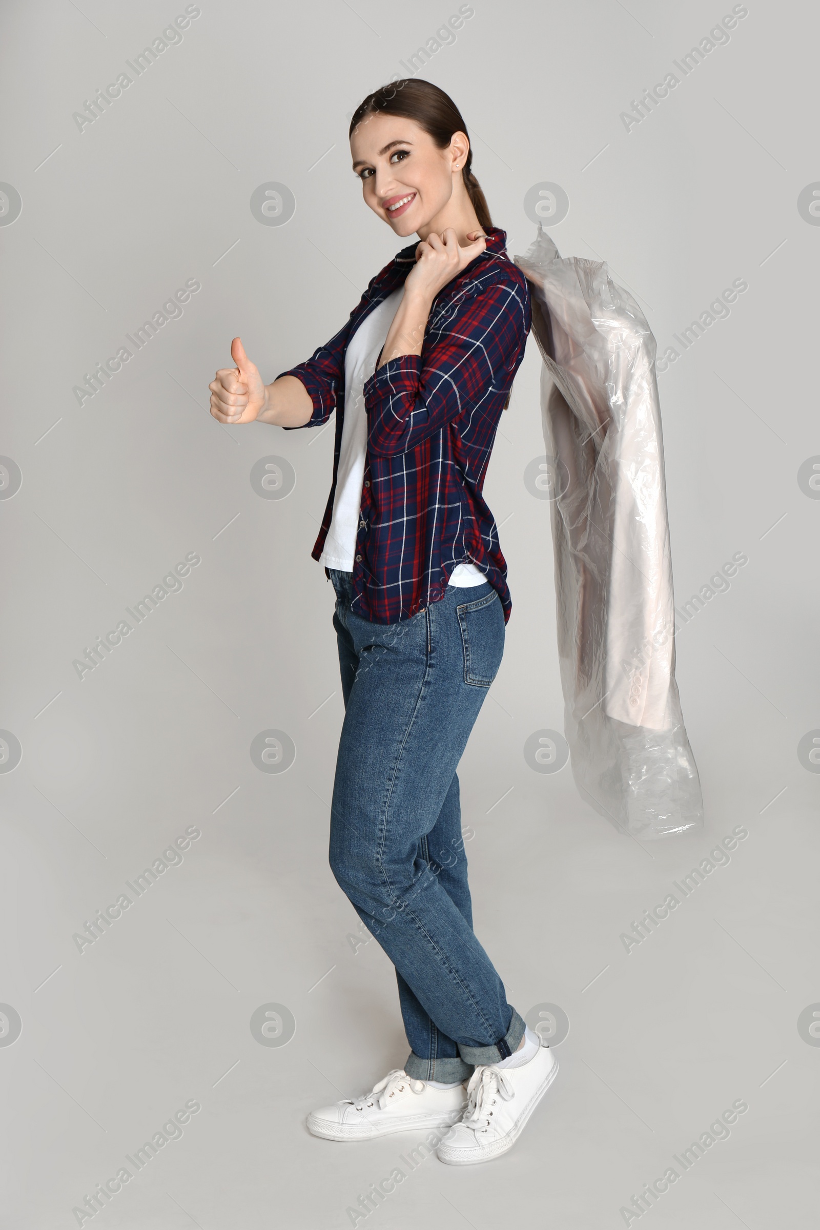 Photo of Young woman holding hanger with jacket on light grey background. Dry-cleaning service