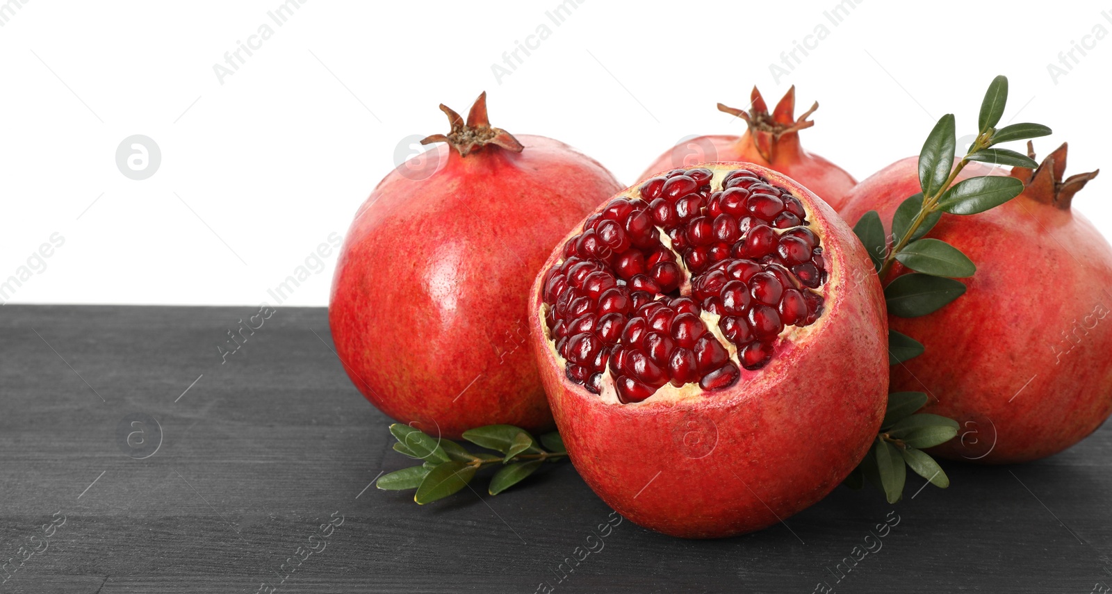 Photo of Fresh pomegranates and green leaves on black wooden table against white background, space for text