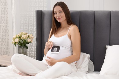 Photo of Pregnant woman with ultrasound picture of baby on bed indoors
