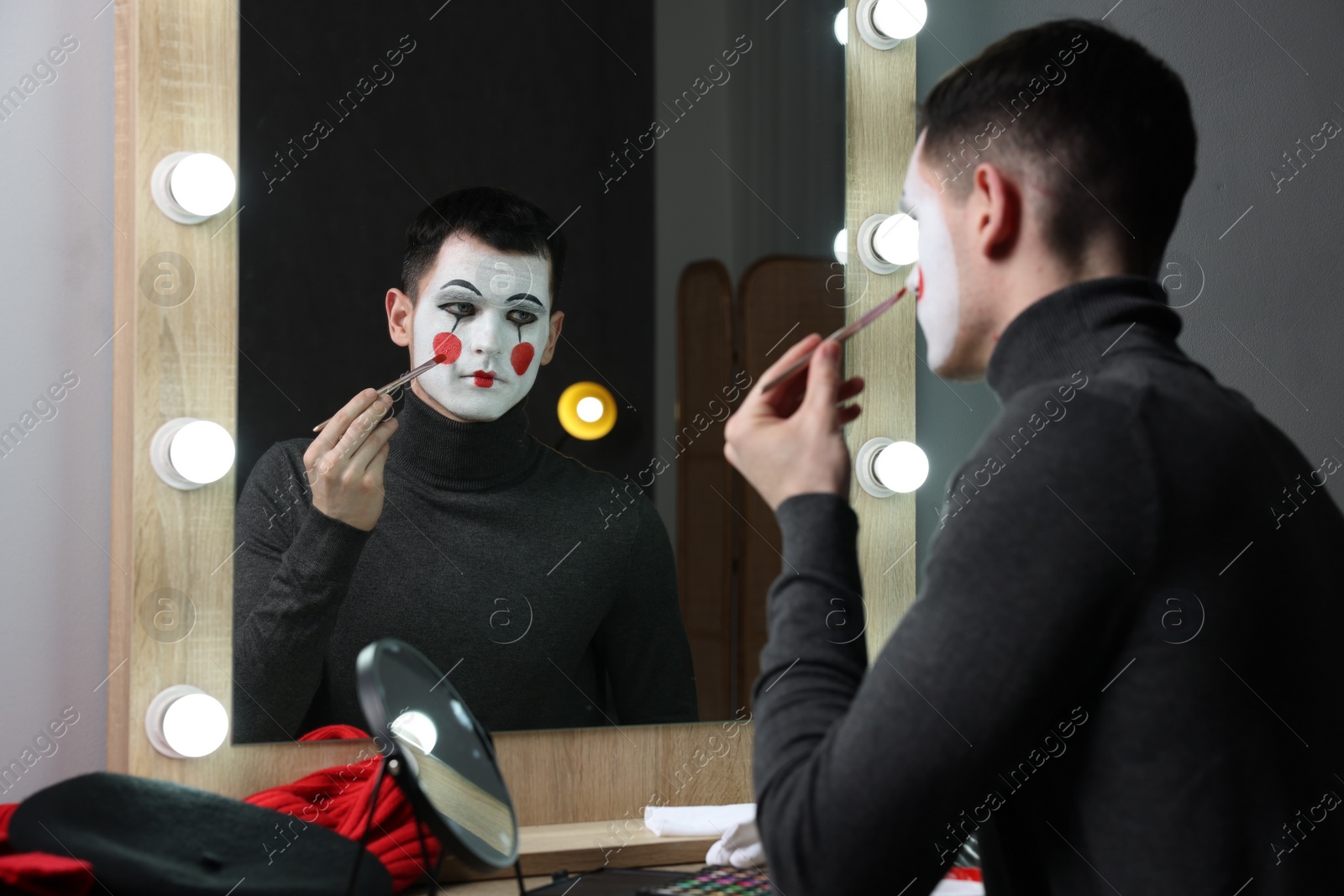 Photo of Young man applying mime makeup near mirror in dressing room