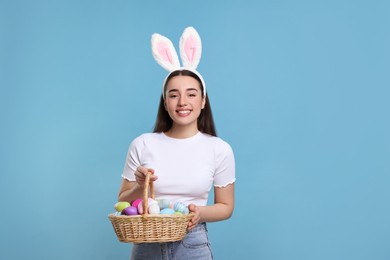 Happy woman in bunny ears headband holding wicker basket of painted Easter eggs on turquoise background