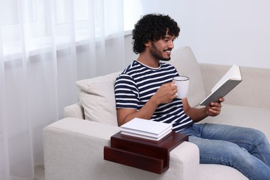 Happy man reading book and holding cup of drink on sofa with wooden armrest table at home