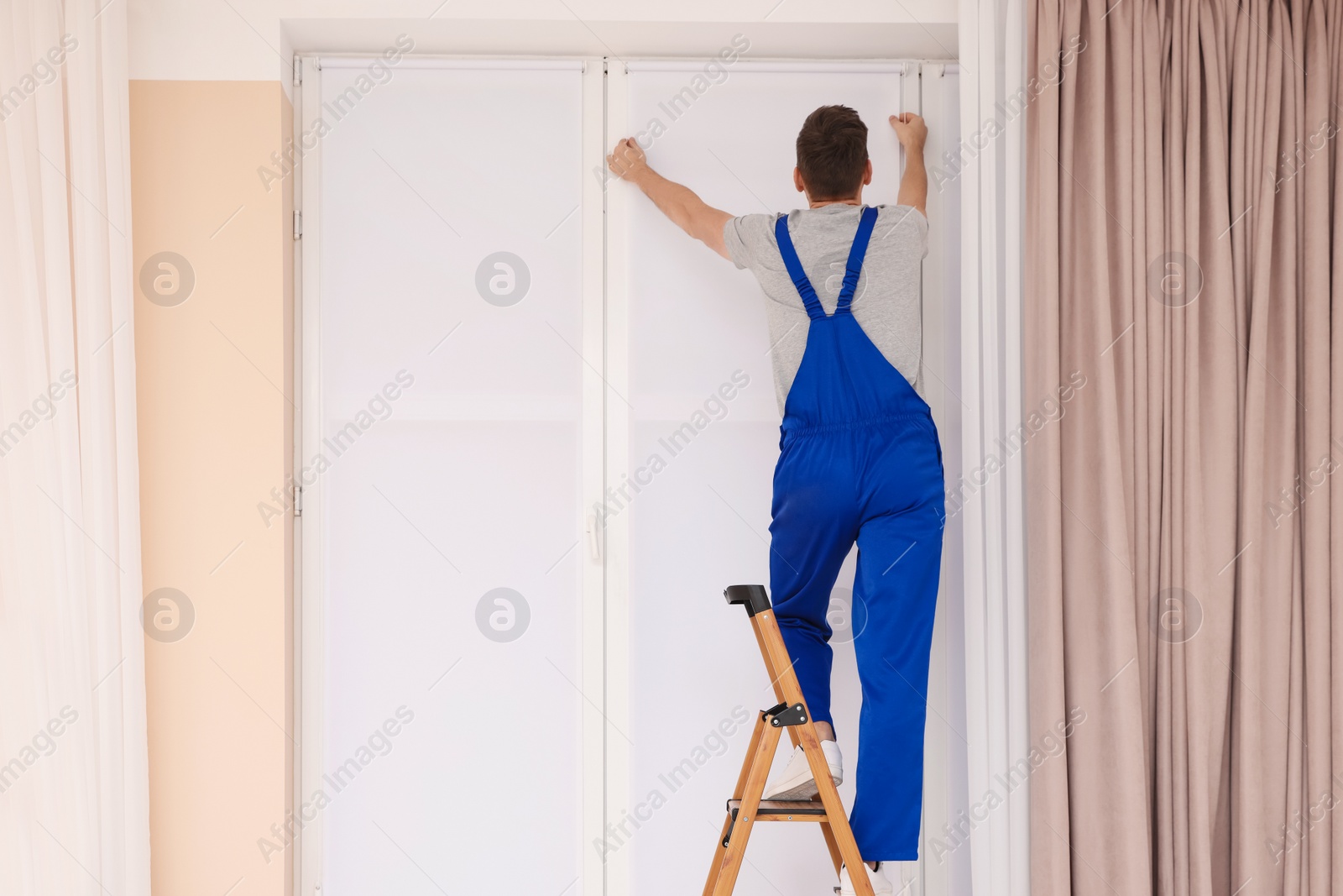 Photo of Worker in uniform installing roller window blind on stepladder indoors, back view