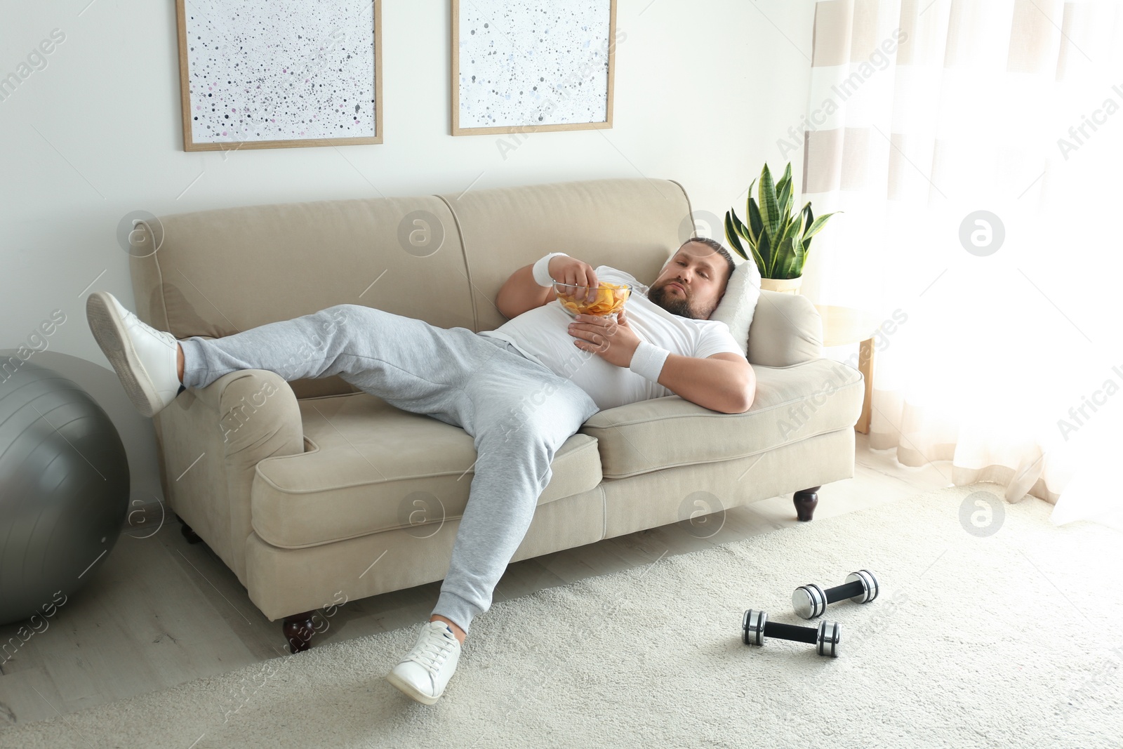 Photo of Lazy overweight man eating chips on sofa at home