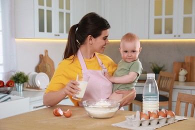 Happy young woman holding her cute little baby while making dough in kitchen