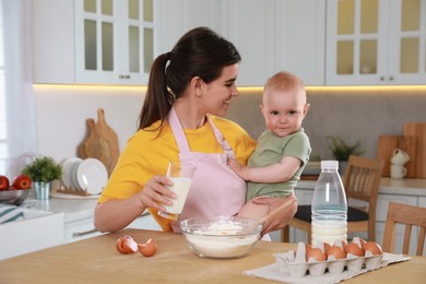 Photo of Happy young woman holding her cute little baby while making dough in kitchen