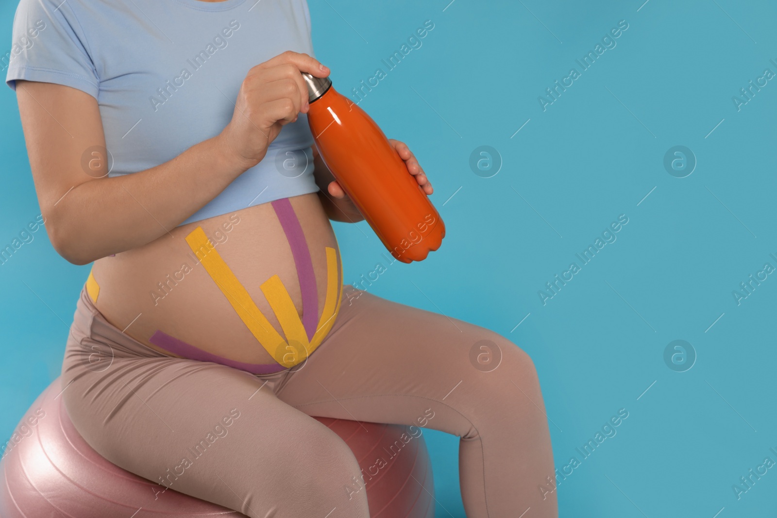 Photo of Pregnant woman with kinesio tapes holding water bottle on fitball against light blue background, closeup. Space for text