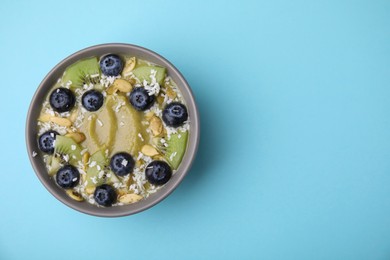 Photo of Bowl of delicious fruit smoothie with fresh blueberries, kiwi slices and coconut flakes on light blue background, top view. Space for text
