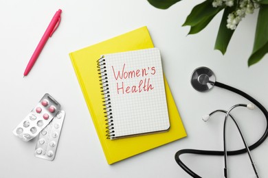 Photo of Notebook with text Women's Health, pills and stethoscope on white table, flat lay