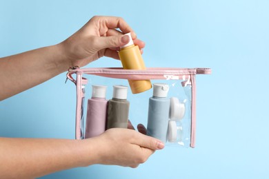 Cosmetic travel kit. Woman putting small bottle with personal care product into plastic bag against light blue background, closeup