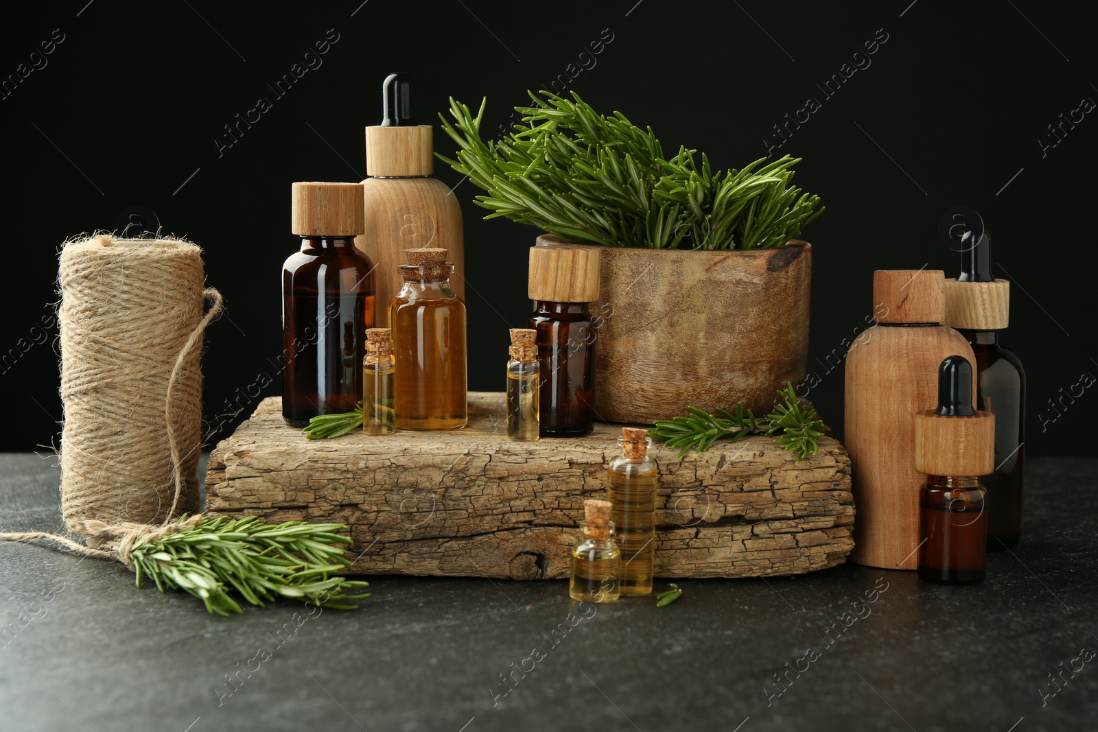 Photo of Essential oils in bottles, rosemary, thread and wood on gray table against black background