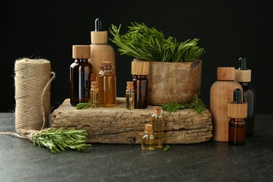 Photo of Essential oils in bottles, rosemary, thread and wood on gray table against black background