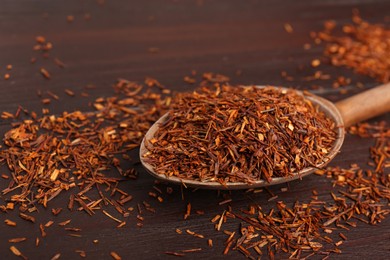 Photo of Spoon with dry rooibos leaves on wooden table, closeup