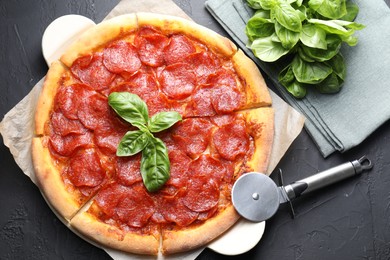 Photo of Tasty pepperoni pizza, basil and cutter on black textured table, flat lay