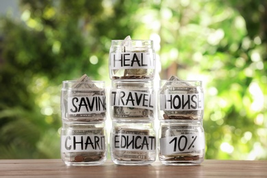 Glass jars with money for different needs on table against blurred background