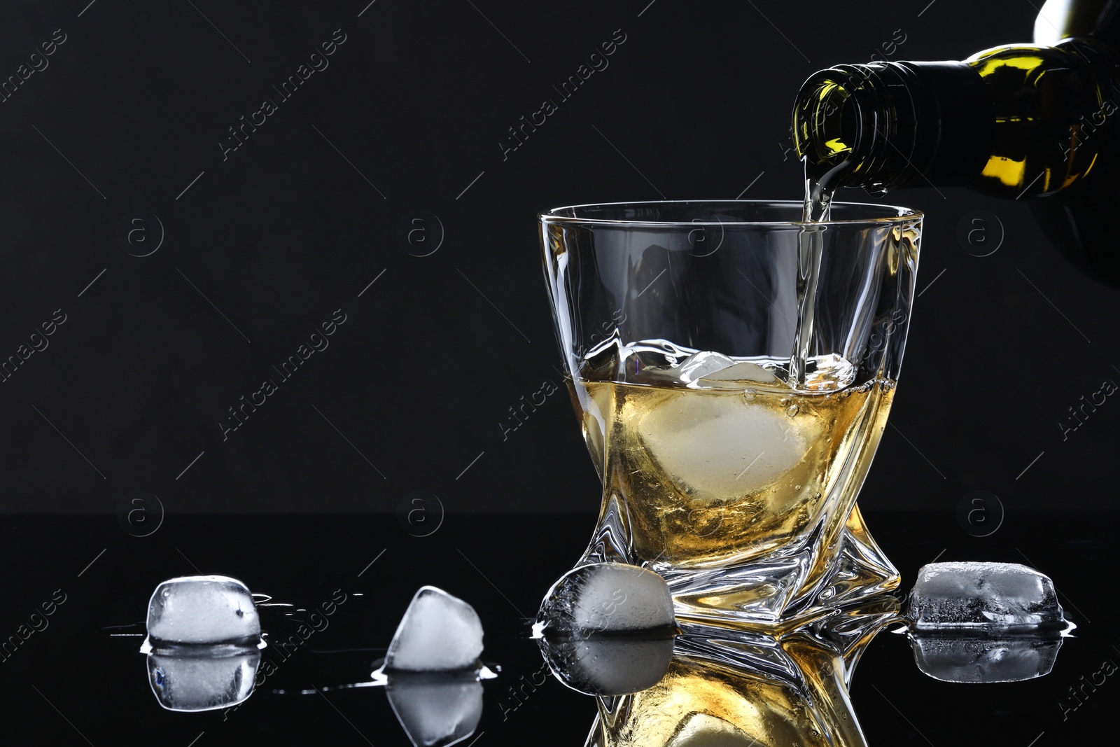 Photo of Pouring tasty whiskey from bottle into glass with ice at mirror table against black background, closeup. Space for text