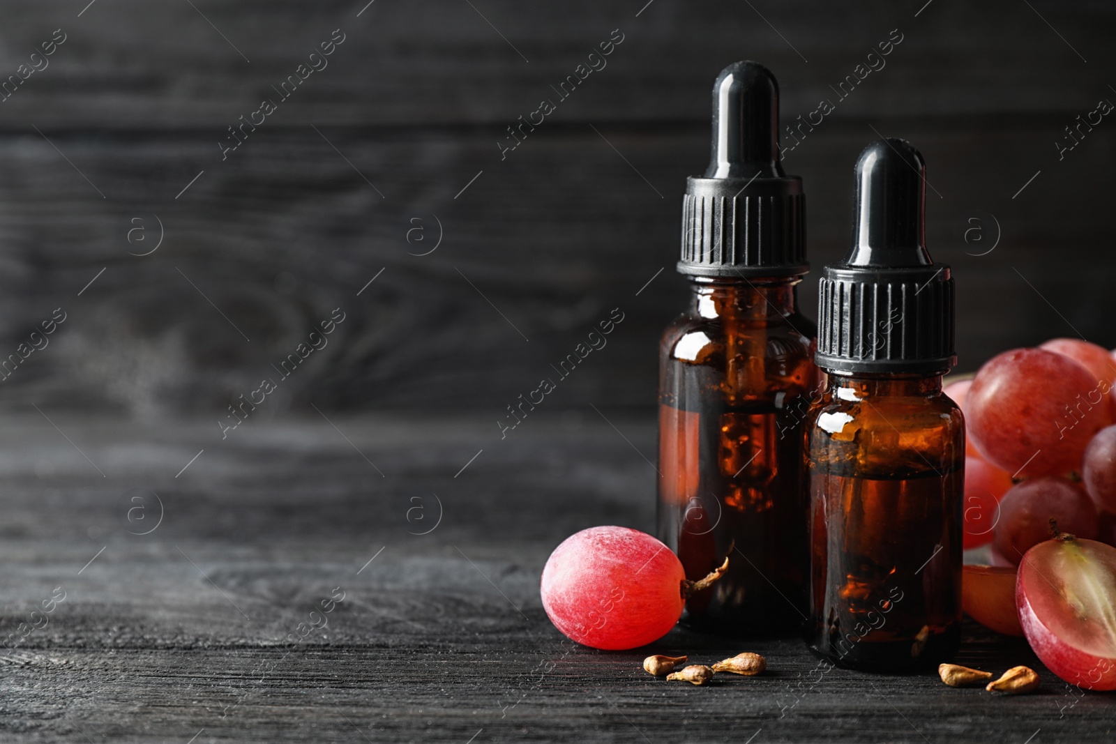 Photo of Organic red grapes, seeds and bottles of natural essential oil on black wooden table. Space for text