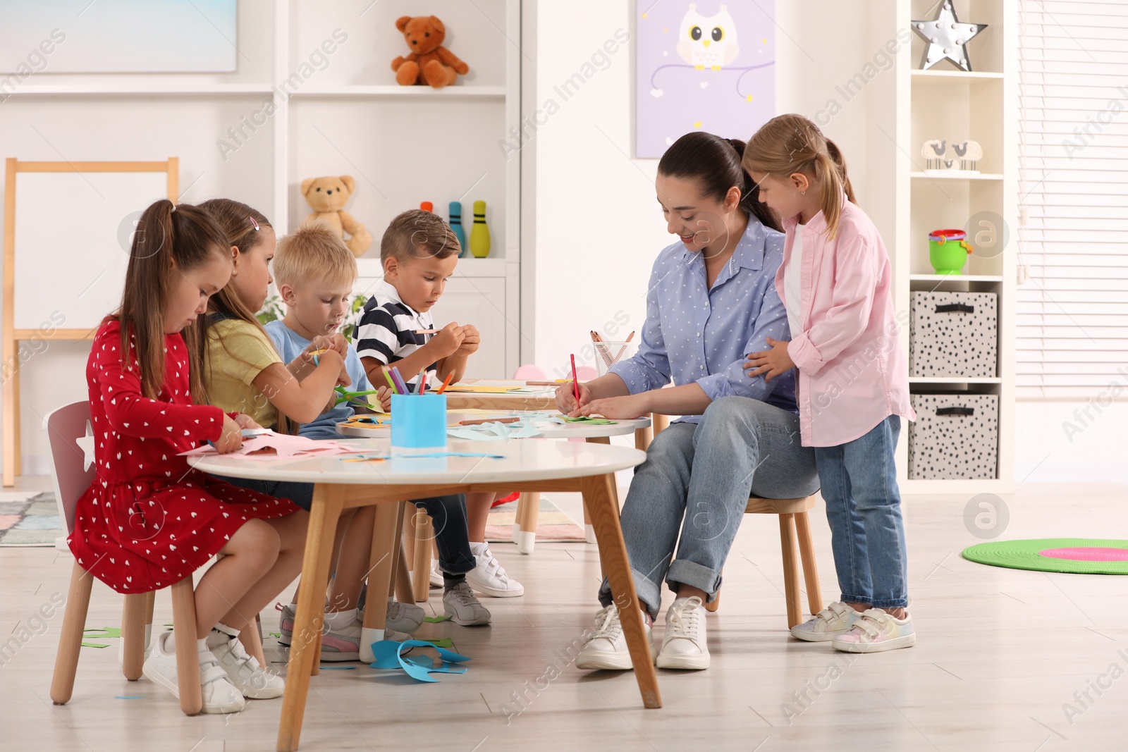 Photo of Nursery teacher and group of cute little children making toys from color paper at desks in kindergarten. Playtime activities