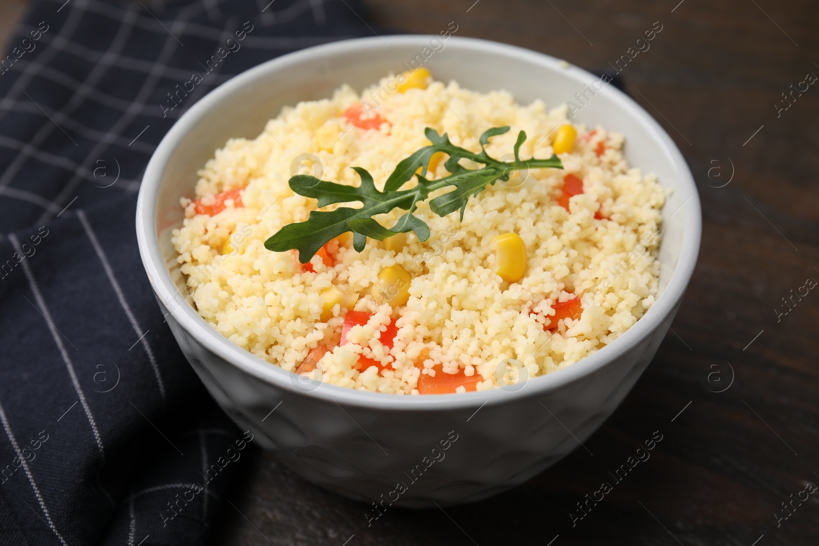 Photo of Tasty couscous with pepper, corn and arugula in bowl on wooden table, closeup