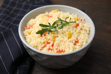 Tasty couscous with pepper, corn and arugula in bowl on wooden table, closeup