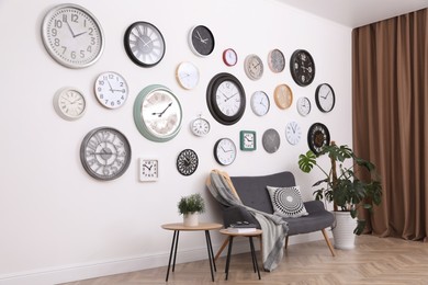 Stylish room interior with comfortable furniture, beautiful houseplant and collection of different clocks on white wall