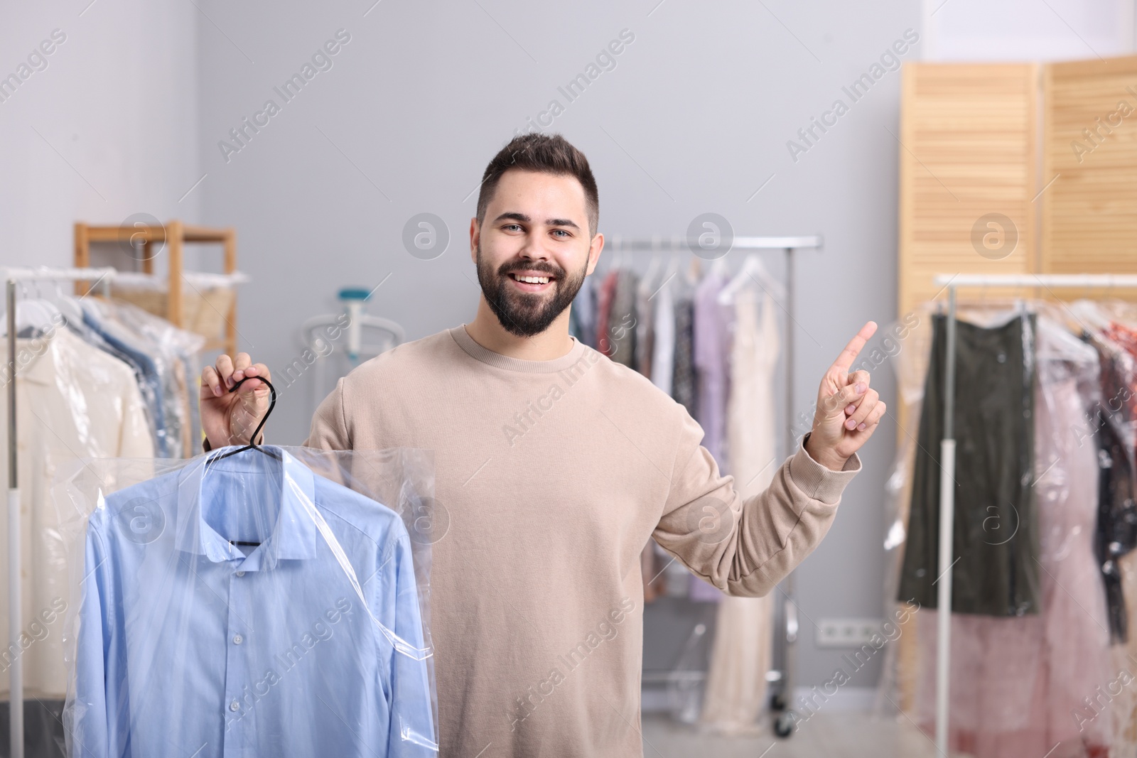 Photo of Dry-cleaning service. Happy man holding hanger with shirt in plastic bag and pointing at something indoors